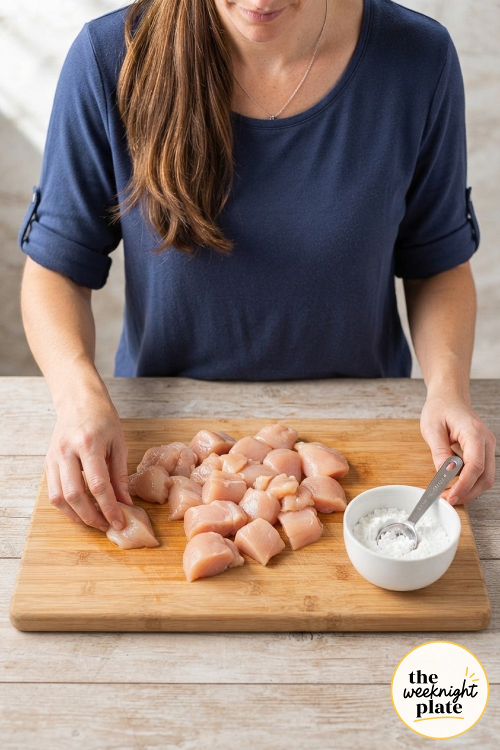 Raw bite-sized chicken pieces on a wooden board beside cornstarch, the first step toward baked orange chicken.